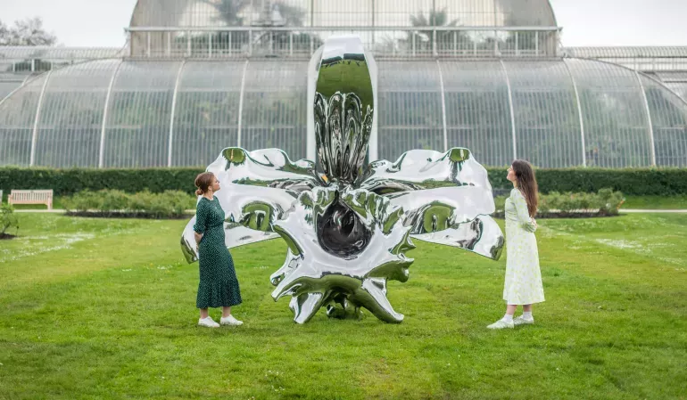 © RBG Kew Two young women stand on either side of a giant shiny metal sculpture of an orchid flower, on a lawn in front of a Victorian glasshouse