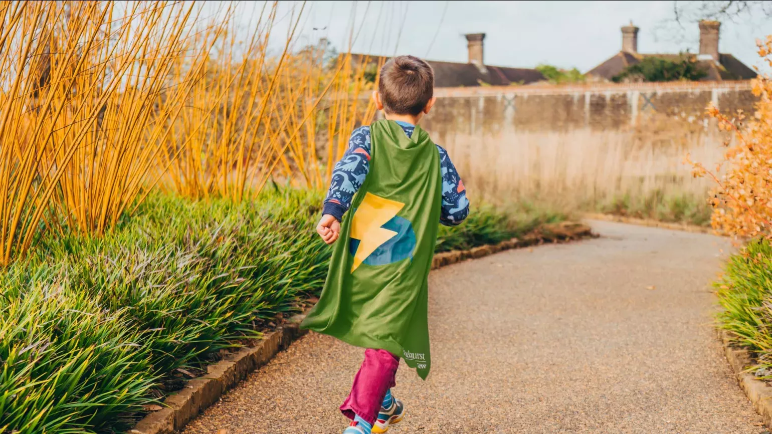 Nature Heroes at Wakehurst, Visual Air © RBG Kew A child runs through the Winter Garden at Wakehurst with a green Nature Heroes cape