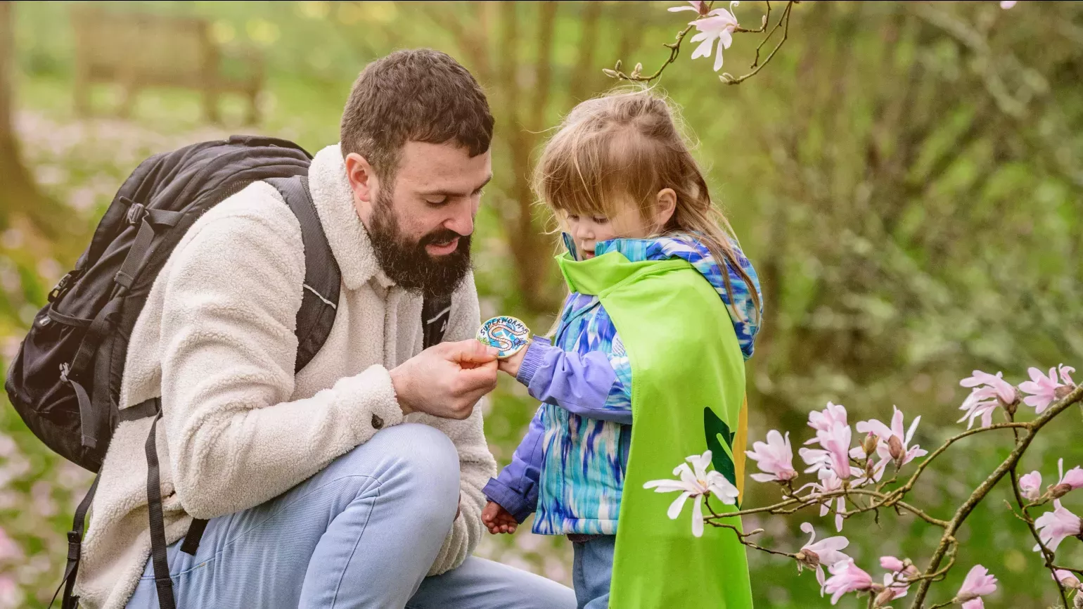 Superworm at Wakehurst © RBG Kew A young girl in a green Nature Heroes cape examines a badge in her hand with her dad, a bearded man in outdoor clothing kneeling next to her. A green park is behind them and pink magnolia blossoms are in the foreground
