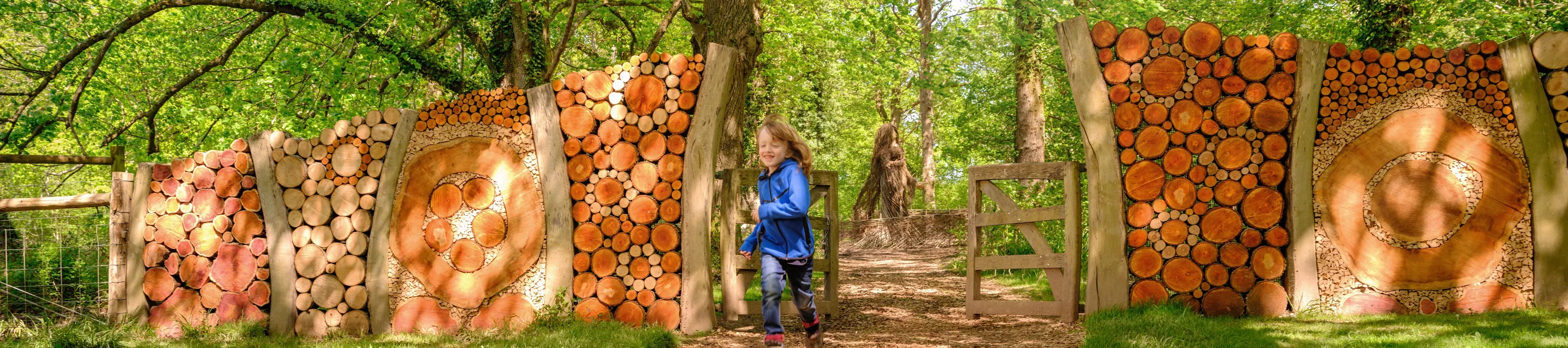 Wild Wood at Wakehurst, Jim Holden © RBG Kew A child runs through a gate in a woodland, made from wooden logs.